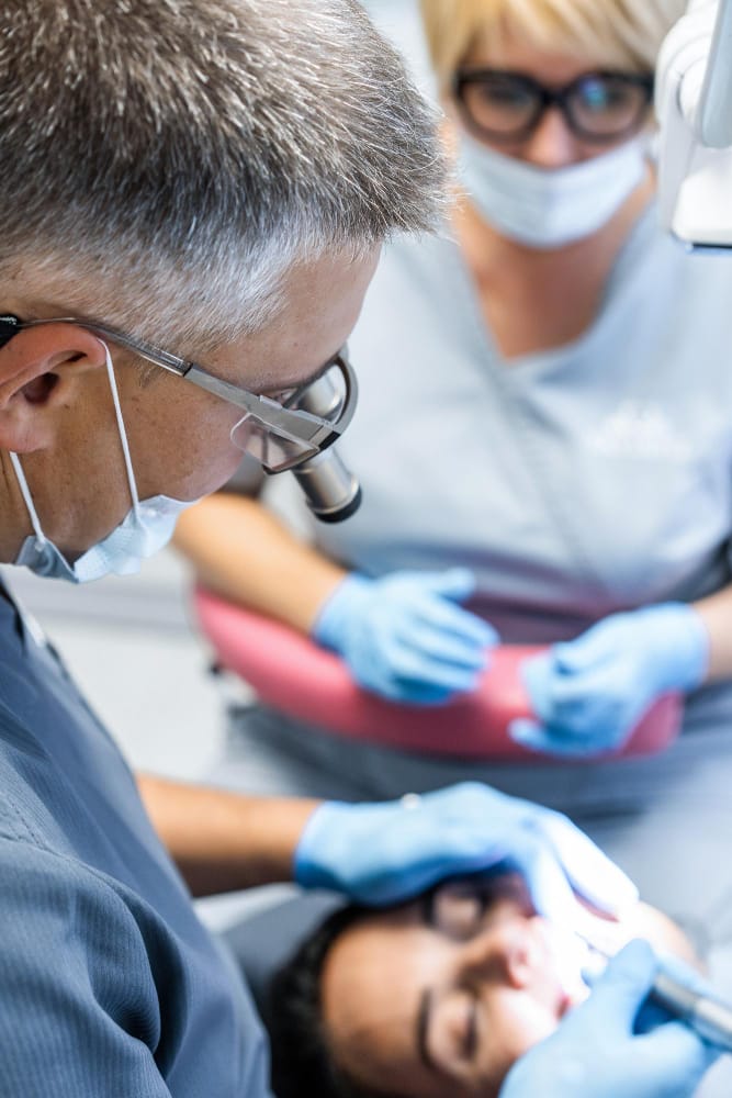 A dental professional wearing magnifying glasses and a mask attending to a patient's mouth, with an assistant in the background, also wearing a mask and gloves.