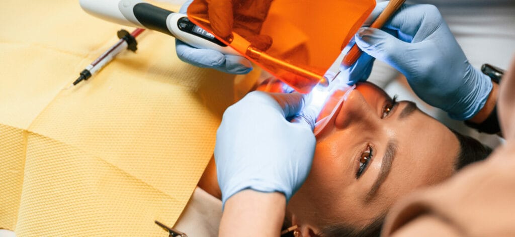 A dentist wearing blue gloves uses dental tools and a curing light on a patient's teeth, while the patient lies back with a protective bib.