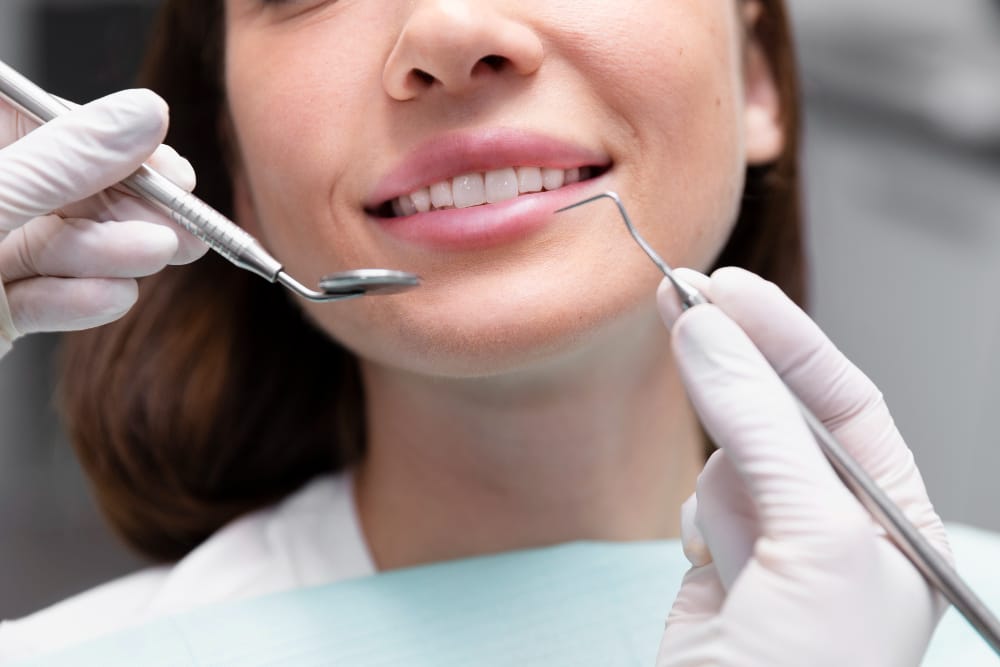 A close-up of a woman at the dentist's office, showing dental tools near her teeth while she smiles.