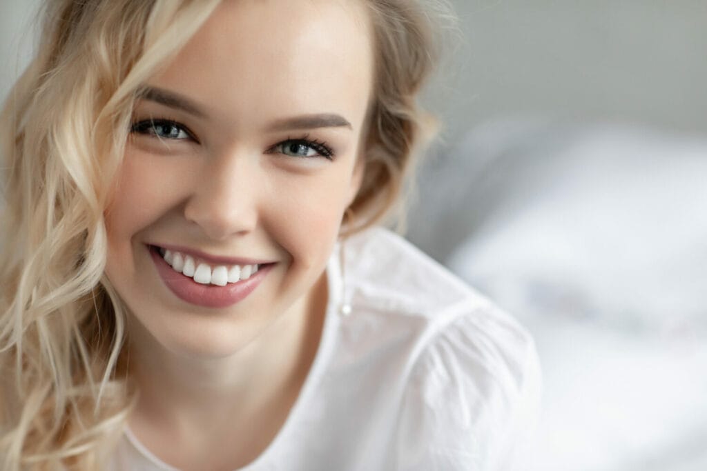 A young woman with blonde hair smiling brightly, showcasing her white teeth, wearing a white top.