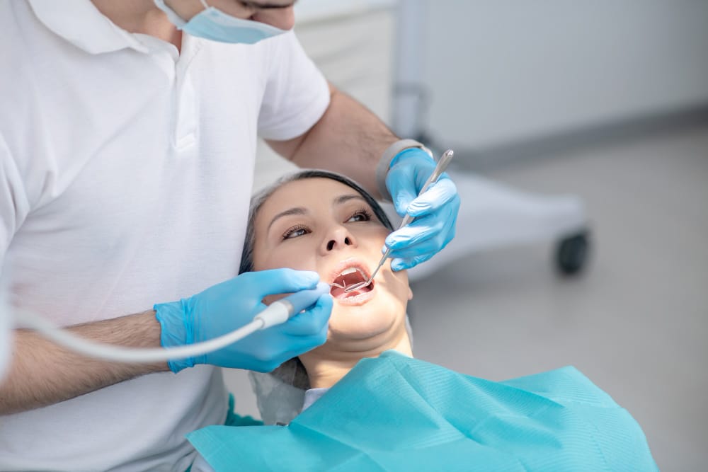 "A dentist wearing protective gear performs a cosmetic dental procedure on a patient sitting in a dental chair. The patient is smiling while the dentist uses a specialized tool to whiten or shape their teeth under a bright examination light.